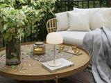 En hyggelig udendørs terrasse med et rundt træbord, en lille lampe, stearinlys, bøger, blomster og en hvid sofa med hynder og et gråt tæppe.