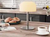 En moderne bordlampe med en rund lampeskærm står på en køkkenbordplade af træ ved siden af en tallerken med kager, en kop kaffe og et åbent magasin.