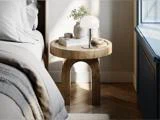 Et rundt natbord af træ med en lampe, en vase og en bog ved siden af en seng med gråt sengetøj, tæt ved et vindue med blå lister, hvor sollyset strømmer ind.
