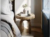 På et rundt natbord af træ står der en hvid lampe, en lille vase med grønne planter, en stak bøger og en dekorativ genstand ved siden af en seng med sengetøj i neutrale farver tæt på et vindue.