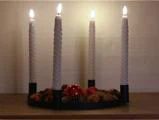 En adventskrans med fire tændte, spiralformede hvide stearinlys, pyntet med fyrrekogler og røde pynteting.