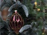 Et facetteret, dybrødt julepynt med guldglitterdetaljer, der hænger på en flokket stedsegrøn gren.