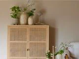 Et skab af lyst træ med døre af rattan, hvorpå der står potteplanter, blomstervaser og en lille lampe.