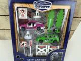 Legetøjsbybilsæt i emballage med en pink og en grøn bil, trafikskilte, træer, kegler og vejafspærringer med påskriften "Caution". På emballagen står der "Little Explorer" og "City Car Set".