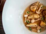 Nærbillede af en gourmetret med grillede rodfrugter, svampe og friske krydderurter i en let bouillon, serveret i en hvid skål.