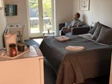Et hyggeligt hotelværelse med en dobbeltseng, en kvinde, der sidder på en sofa, et tv, et skrivebord og et tekøkken med kaffeudstyr; sollyset strømmer ind gennem glasdørene.