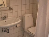 Et lille badeværelse med et hvidt toilet, en væghængt håndvask, flisebelagte vægge, en spejlhylde og et hvidt bruseforhæng med små prikker.