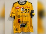 En gul sportstrøje med sorte striber, nummer 22 og forskellige sponsorlogoer, der hænger på en bøjle mod en hvid struktureret væg.