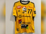 En gul sportstrøje med sorte detaljer, nummer 77 og forskellige sponsorlogoer, der hænger på en sort bøjle mod en hvid struktureret væg.