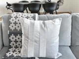 En lysegrå pude hviler på en grå sofa med et mønstret tæppe. I baggrunden ses et sort spisebord med matchende stole.
