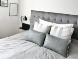 En pænt redt seng med grå og hvide puder, et tuftet gråt hovedgærde og to indrammede citater på hvide vægge med en sort vægmonteret lampe ved siden af sengen.