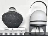En sort tekstureret vase med en glaskugle på toppen står på stablede bøger ved siden af en moderne hvid lampe med en sort ramme, alt sammen mod en hvid væg.