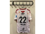 En hvid sportstrøje med røde detaljer hænger på en vægkrog. På ryggen ses nummeret 22 og navnet NOLSØE samt forskellige sponsorlogoer.