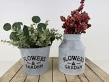 To metalkrukker med påskriften FLOWERS & GARDEN står på en træplade; den ene indeholder grønne blade, den anden røde bærlignende stængler.