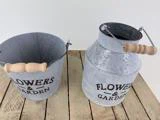 To metalbeholdere med træhåndtag mærket FLOWERS & GARDEN står på en træoverflade; den ene er en spand, den anden er en kande.