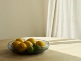 En glasskål fyldt med citroner og limefrugter står på et lyst træbord, hvor sollyset strømmer ind gennem et vindue med hvide gardiner.