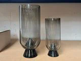 To gråtonede glasvaser med sorte, koniske fødder står side om side på en lysebrun overflade foran en ensfarvet baggrund.
