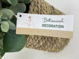 Et mærke med teksten Botanical Decoration sidder på en flettet kurv med grønne kunstige blade, der er delvist synlige omkring den.