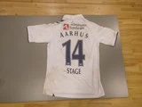 En hvid fodboldtrøje med AARHUS og nummer 14 trykt på ryggen sammen med et sponsorlogo og navnet STAGE. Trøjen har nogle snavsede mærker og ligger på et bord.