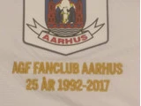 Et hvidt stofmærke med Aarhus' byvåben over teksten AGF FANCLUB AARHUS 25 ÅR 1992-2017 broderet med guldbogstaver.