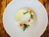 En hvid dessert, muligvis sorbet, serveret med cremet sauce, spiselige blomster og grønne krydderurter på en struktureret hvid tallerken på et rustikt træbord.