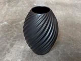 En mat sort, oval vase med en spiralformet, riflet overflade står på en grå betonoverflade.