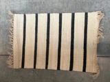 Et rektangulært vævet tæppe med beige og sorte lodrette striber og frynser langs to kanter, der ligger på et gråt betongulv.