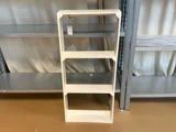 En enkel hvid træreol med fire hylder står på et brunt gulv mellem metalreoler i en butik.