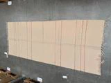 Et rektangulært beige tæppe med tynde orange og lyserøde lodrette striber og frynser langs kanterne ligger på et gråt betongulv.