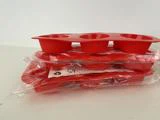 Tre røde silikone-muffinsforme, hvoraf to stadig er pakket ind i plastik, er stablet på en hvid flade mod en lys struktureret væg.