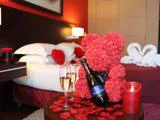Et romantisk hotelværelse med en seng dekoreret med rosenblade og hjertepuder, et bord med champagne, glas, et stearinlys og en bamse lavet af røde roser.
