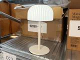 En lille hvid bordlampe med en kuppelformet, riflet lampeskærm står på en metalhylde tæt ved nogle papkasser i en butik eller et lager.