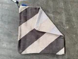 Et foldet, strikket tæppe med brede diagonale striber i sort og beige ligger på en grå betonoverflade.