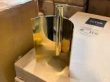 Et skinnende, guldfarvet metalgenstand med et cylindrisk rør og buede sider står på en papkasse blandt andre kasser.