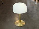 En moderne bordlampe med fod og stamme af messing, kronet af en hvid, kuppelformet lampeskærm, står på et poleret betongulv.