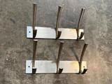 To metalliske vægophængte frakkestativer med tre kroge hver, placeret parallelt på en struktureret, solbeskinnet overflade.