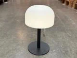 En lille bordlampe med en mat hvid, kuppelformet lampeskærm, sort fod og trækæde, der står på et gråt gulv i et lagerlokale.