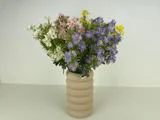 En beige, riflet vase med en buket hvide, pink, gule og lilla vilde blomster på en almindelig hvid baggrund.