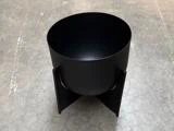 En mat sort, cylindrisk blomsterkrukke, der står på et krydsformet metalstativ.
