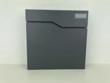 En moderne, mørkegrå postkasse med et geometrisk design står mod en lys struktureret væg. En rektangulær etiketholder er synlig i øverste højre hjørne.