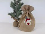 En lille gavepose i jute med et rensdyr med nissehue står ved siden af et lille kunstigt juletræ pakket ind i jute.