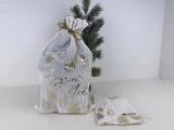 En hvid gavepose med guldpynt og Joyeux Noël skrevet på ligger ved siden af et sammenfoldet hvidt klæde og et lille kunstigt fyrretræ.