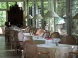 En række elegante borde med hvide duge og røde servietter står i en solbeskinnet restaurant med store vinduer og kurvestole.