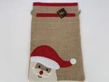 En gavepose af jute med en rød prikket kant og en applikation af julemandens ansigt med en rød hue i nederste venstre hjørne.