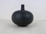 En lille, rund, mørkegrå keramikvase med smal hals, placeret på en lys overflade mod en almindelig hvid baggrund.