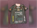 Grøn sportstrøje med nummer 35, flere sponsorlogoer og teksten »DET FAGLIGE HUS«. Trøjen er udstillet på en lilla, struktureret overflade.