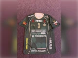 En grøn sportstrøje med nummer 3, forskellige sponsorlogoer og hvid kant ligger fladt på en lilla mønstret overflade.
