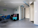 Moderne, minimalistisk opholdsrum med en sort sofa, et spisebord med blå stole, en stige op til en hems og hvide døre; lyse vægge og gulv.