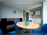 Et lille, moderne spiseområde med et rundt bord, blå stole, to grønne glasvaser, en sort og grå sofa samt et kompakt, hvidt køkken i baggrunden.