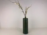 En høj, mørkegrøn keramikvase holder to lange grene med små hvide blomster mod en almindelig hvid baggrund.